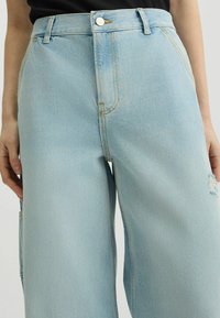 Pantalones anchos de mezclilla azul claro con cintura alta, que cuentan con cierre de botón y un ligero desgaste a lo largo del costado.