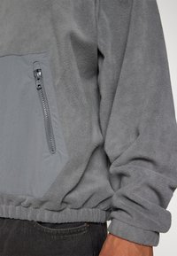 Šedý fleecový svetr s texturovaným tělem, hladkým šedým kapsou se zipovým uzavíráním a elastickými manžetami a lemem.