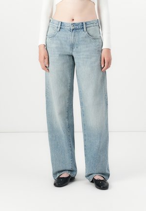 Frau trägt hellblaue Jeans mit weitem Bein, ein weißes, kurzes langärmeliges Oberteil und schwarze Ballerinas und steht vor einem weißen Hintergrund.