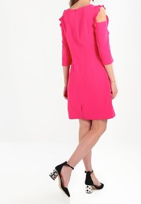 Robe rose avec manches trois-quarts et épaules découpées. Dotée de volants et d'une fermeture éclair cachée dans le dos. Talons noirs avec talons carrés à motif.