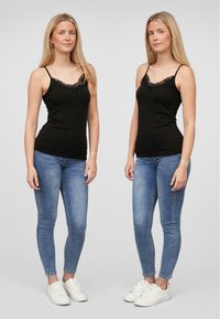 Svart topp med kant i blonder iført blå smale jeans. Modellen står i to poseringer, med tynne stropper og en tettsittende silhuett. Hvite sneakers.
