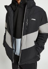 Giubbotto imbottito con pannelli neri, grigi e blu chiaro, chiusura con zip, cappuccio e logo sul petto. Texture morbida e trapuntata per il calore.
