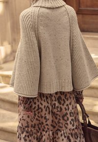 Personne portant un pull beige en maille  à manches larges sur une robe à imprimé léopard, tenant un sac en cuir marron, debout sur des marches en pierre.