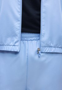 Μαλακό γαλάζιο μπουφάν με φερμουάρ και παντελόνι με ελαστική μέση που ταιριάζει, πάνω από μαύρο πουκάμισο, δείχνοντας τη λεπτομέρεια του φερμουάρ και την υφή του υφάσματος.