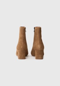 Botas de ante marrón hasta el tobillo con tacones gruesos y cremallera lateral. Textura suave, líneas limpias y diseño de punta redonda.
