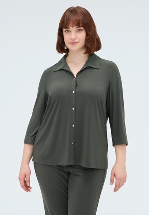 Femme portant un chemisier vert foncé boutonné avec des manches trois quarts et un pantalon assorti, debout devant un fond clair uni.