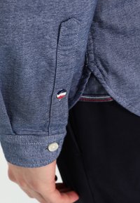 Camisa de botones con una tela texturizada de color azul oscuro, que presenta un botón blanco en el puño y un pequeño logo bordado en rojo, blanco y azul.
