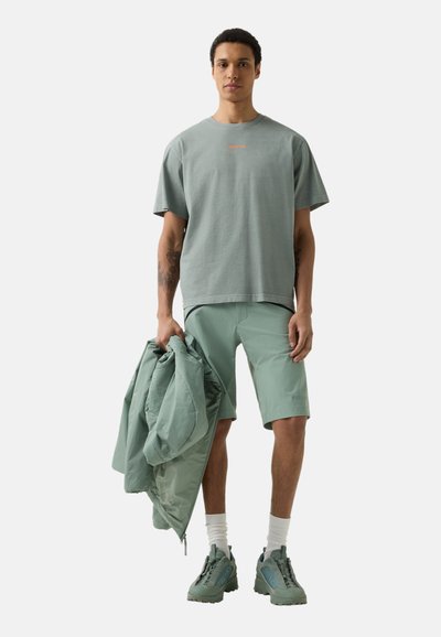 Mladý muž v zelených šortkách, nadměrně velkém tričku, turistických botách a držící zelenou bundu na jednobarevném světle pozadí.