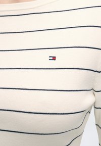 Chemise blanche en maille à manches longues avec de fines rayures horizontales noires et un petit logo rectangulaire rouge, blanc et bleu sur la poitrine.