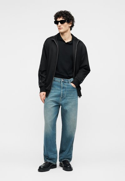 Νέος άνδρας που φοράει μαύρα γυαλιά ηλίου, μαύρο μπουφάν με φερμουάρ, μαύρο πουκάμισο, ξεθωριασμένο μπλε τζιν και μαύρα παπούτσια, στέκεται με το ένα χέρι στην τσέπη.