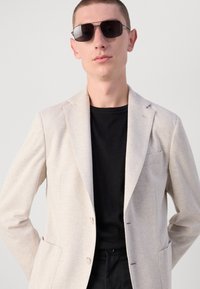 Blazer a quadri beige chiaro con una tasca singola, abbinato a una t-shirt nera. La modella indossa occhiali da sole rettangolari scuri.
