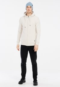 Felpa zip beige chiaro con cappuccio regolabile, polsini e orlo a coste. Abbinata a jeans neri e a un berretto di maglia azzurro chiaro. Scarpe nere.