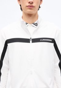 Νεαρός με λευκό μπουφάν με φερμουάρ και μαύρη ρίγα, με το λογότυπο «J.LINDEBERG» στο στήθος, και λευκό πουκάμισο με μαύρο σχεδιασμένο γιακά από κάτω.