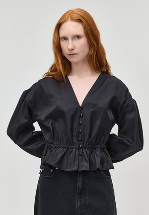 Žena s dlhými červenými vlasmi, oblečená v čiernej blúzke so zapínaním na gombíky a sťahovaním v páse, v kombinácii s tmavými džínami s vysokým pásom, pózujúca s rukami za chrbtom.