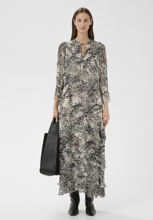 Femme portant une longue robe à motif noir et blanc, tenant un grand sac à main noir et portant des bottines noires sur un fond blanc.