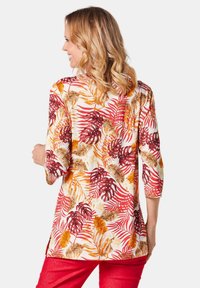 Blütenmuster-Tunika in Creme mit roten, orangefarbenen und braunen Blättern. Mit drei Viertel langen Ärmeln und seitlichen Schlitzen für mehr Bewegungsfreiheit.