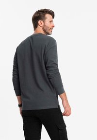 Mann mit kurzem braunen Haar und Bart, der ein dunkelgraues Langarmshirt und schwarze Cargo-Hosen trägt, seitlich vor weißem Hintergrund stehend.