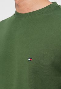 Közelkép egy személyről, aki zöld kereknyakú pólót visel, amin egy kicsi piros, fehér és kék téglalap alakú logó látható a mellkasán.