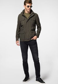Oliwkowa kurtka z kapturem, z wieloma przednimi kieszeniami, na zamek błyskawiczny i z guzikami, połączona z czarnymi przetartymi dżinsami i czarnymi trampkami.