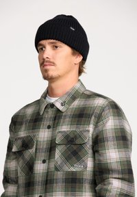 Karierter Flanellhemd in Grün, Schwarz und Weiß mit geknöpften Brusttaschen. Schwarze Rippmütze. Modell mit hellem Bart.