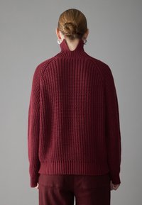 Burgunderfarbener Strickpullover mit hohem Kragen, strukturiertem Maschenmuster und gerippten Säumen, kombiniert mit passenden burgunderfarbenen Hosen.