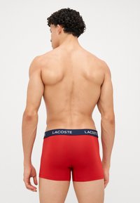 Boxer shorts vermelhos feitos de material suave, com um cós azul-marinho contendo a marca "LACOSTE" em branco. Design de perna curta.