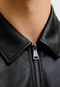 Schwarze Lederjacke mit spitzem Kragen und silbernen Reißverschlussdetails. Glatte Textur mit Nahaufnahme des Reißverschlussbereichs.