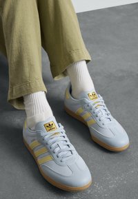 Henkilö, joka käyttää vaaleansinisia ja keltaisia Adidas Samba -tennareita valkoisten sukkien kanssa oliivinvihreiden housujen päällä harmaalla lattialla.