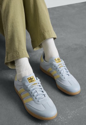 Person som har på seg lys blå og gul Adidas Samba-sneakers med hvite sokker og olivengrønne bukser på grått gulv.