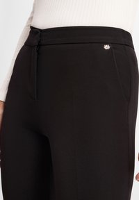 Pantaloni neri su misura con una texture liscia, caratterizzati da un piccolo logo metallico e una chiusura con cerniera laterale.