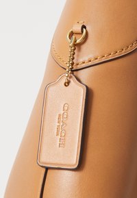 Handtas van kastanjebruin leer met een gladde textuur, voorzien van gouden metalen accenten, een hanglabel met het uitgesneden opschrift "COACH NEW YORK" en contrasterende steekdetails.