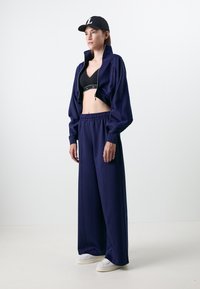 Casaco cropped azul-marinho com gola alta, combinado com um soutien desportivo preto e calças largas azul-marinho. Ténis brancos completam o visual.