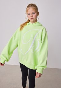 Neon zöld túlméretezett pulóver, fehér logóval, nagy kapucnival és széles ujjakkal, fekete leggingsszel és fehér cipővel párosítva.