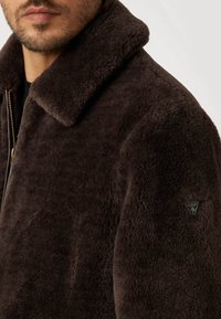 Giubbotto in shearling marrone con ampio colletto, dotato di cerniera argentata e piccolo logo nero sul lato sinistro della spalla. Texture morbida.