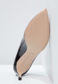 Černé lakované lodičky na vysokém podpatku s špičatou špičkou, béžovou podrážkou a označením velikosti 37, nakloněné proti bílé zdi.