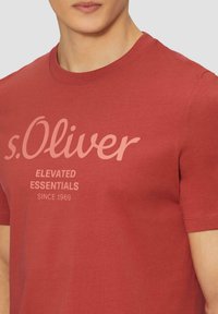Rotes Baumwoll-T-Shirt mit kurzen Ärmeln, das ein markantes hellrosa Schriftlogo "s.Oliver" sowie zusätzlichen Text darunter zeigt. Glatte Textur.