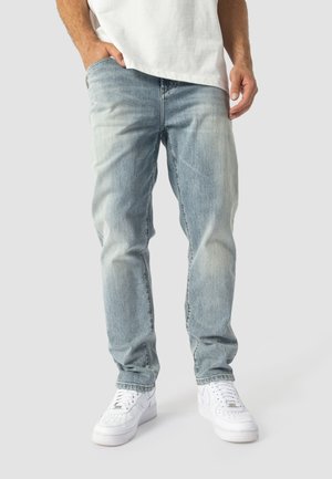 Mann trägt hellblaue ausgewaschene Jeans, weiße Sneakers und ein weißes Hemd mit einer Hand in der Tasche vor einfarbigem Hintergrund.