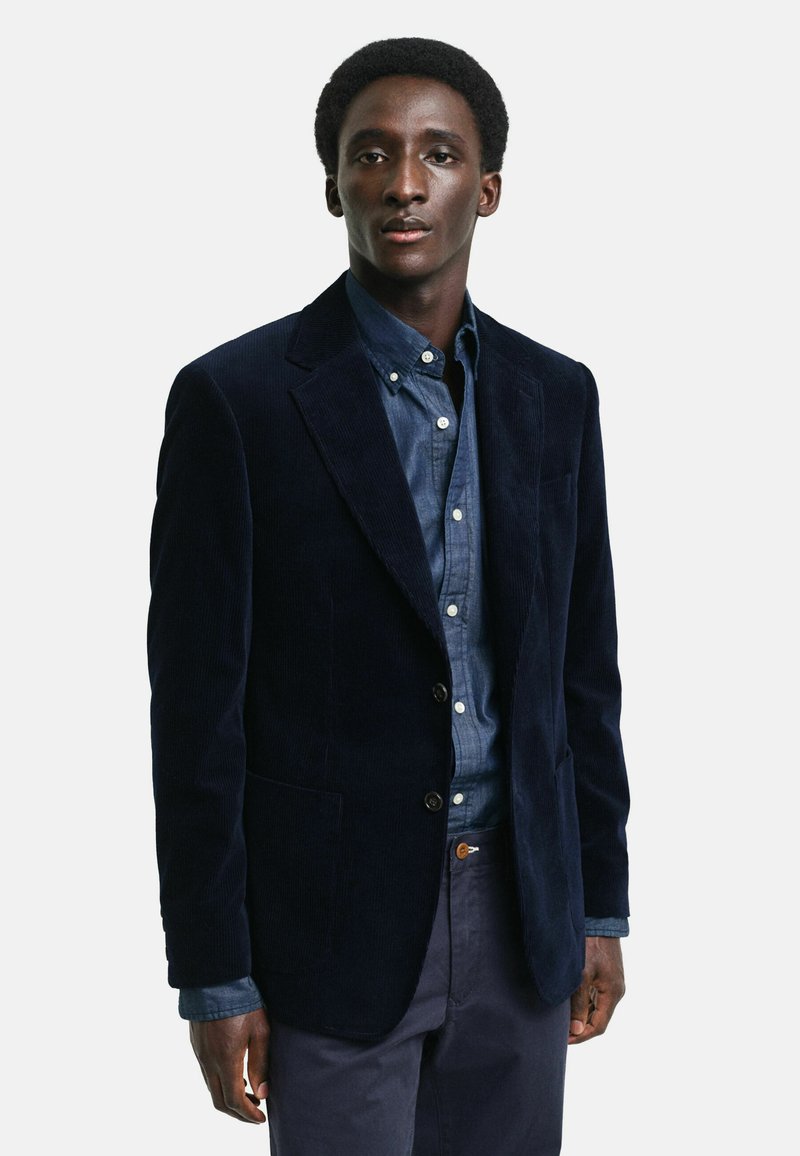 Marineblauer Cord-Blazer mit Revers, zwei Knöpfen und vorderen Taschen, getragen über einem Jeanshemd. Glatte Textur mit vertikaler Rippung.