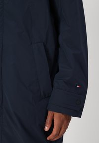 Navy plava padana jakna s čvrstom teksturom, s manžetama na kopčanje i bočnom džepom. Mali logo na rukavu.