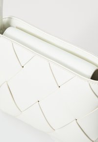 Biała pleciona torebka z teksturowanym, splatanym wzorem. Gładki materiał, prostokątny kształt, z wąskim otworem na górze.