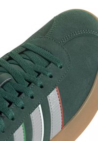 Baskets en daim vert foncé avec des lacets assortis, des bandes latérales blanches et rouges, et une semelle en caoutchouc marron clair.