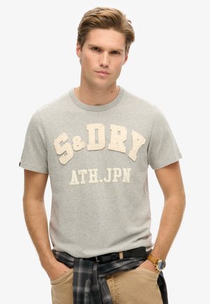 Camiseta gris de manga corta con letras texturizadas en beige "S&D" y "ATH.JPN." Tejido liso; el modelo lleva un cinturón negro y una camisa de cuadros atada a la cintura.