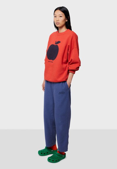 Rød sweatshirt med sort æblegrafik, parret med løse blå bukser og grønne klipklappere. Blødt bomuldsstof, afslappet pasform, casual look.