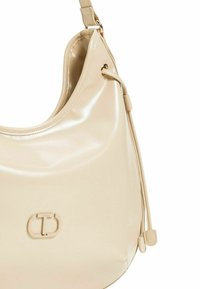 Borsa beige realizzata in materiale lucido. Caratterizzata da una forma arrotondata, una sola tracolla e un accento con logo al centro. Texture liscia.