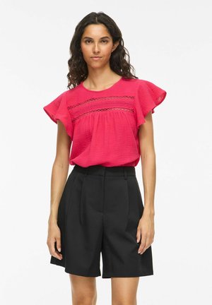 Femme aux longs cheveux bouclés portant un chemisier rose vif avec des détails en dentelle et un short noir taille haute, debout devant un fond blanc.