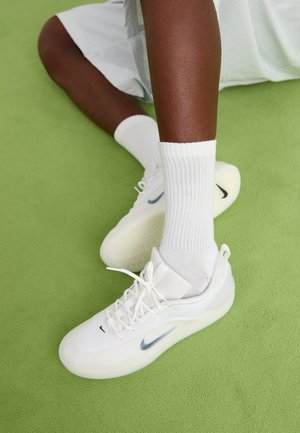 Sneakers blancs Nike avec un dessus lisse, des lacets et une semelle translucide présentent un petit logo noir. Portés avec des chaussettes blanches à côtes. Fond vert.