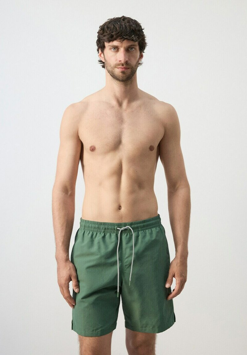 Muž bez košele s krátkymi tmavými vlasmi, oblečený v zelených plavkách so sťahovacou šnúrkou, stojaci pred jednoliatym svetlosivým pozadím, čelí dopredu.