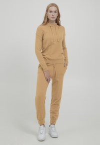 Sennepsgul loungewear-sæt bestående af en hætte-sweater med snore og snævre bukser med sidelommer, parret med hvide sneakers.