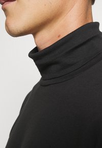 Camisa negra de cuello alto hecha de tela suave con un diseño ajustado, que presenta un alto cuello y una textura lisa. Vista lateral en primer plano.