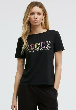 Blonde woman wearing a black T-shirt with colorful "SOCCX" text and cursive "Reg Trademark," standing with one hand in pocket on a plain background.